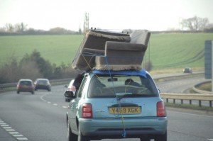 Two sofas are perched on the roof of a small car and the passenger uses his left hand to secure them